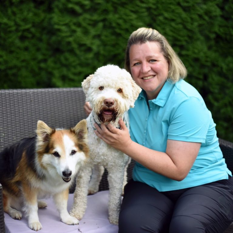 Eine lächelnde Tierphysiotherapeutin mit einem weissen und einem braun-weissen Hund auf einen Bank im grünen