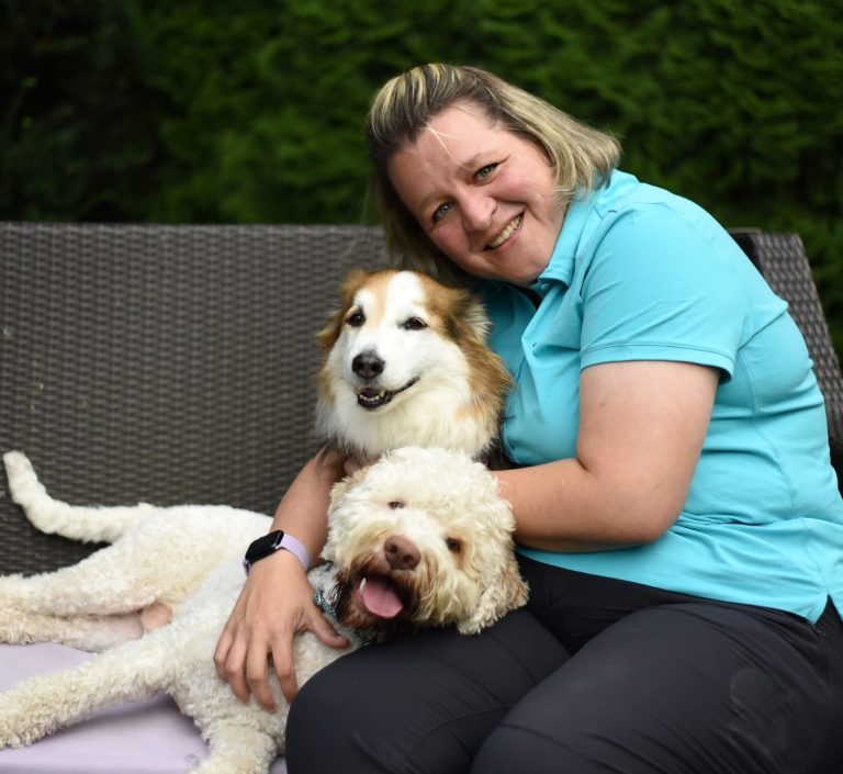 Tierphysiotherapeutin mit Zwei Hunden, die sie liebevoll umarmt, auf einer Bank im Garten.