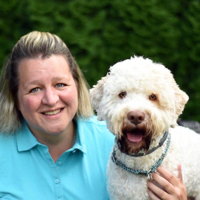 Eine Tierphysiotherapeutin lächelt neben einem weißen Hund mit lockigem Fell.
